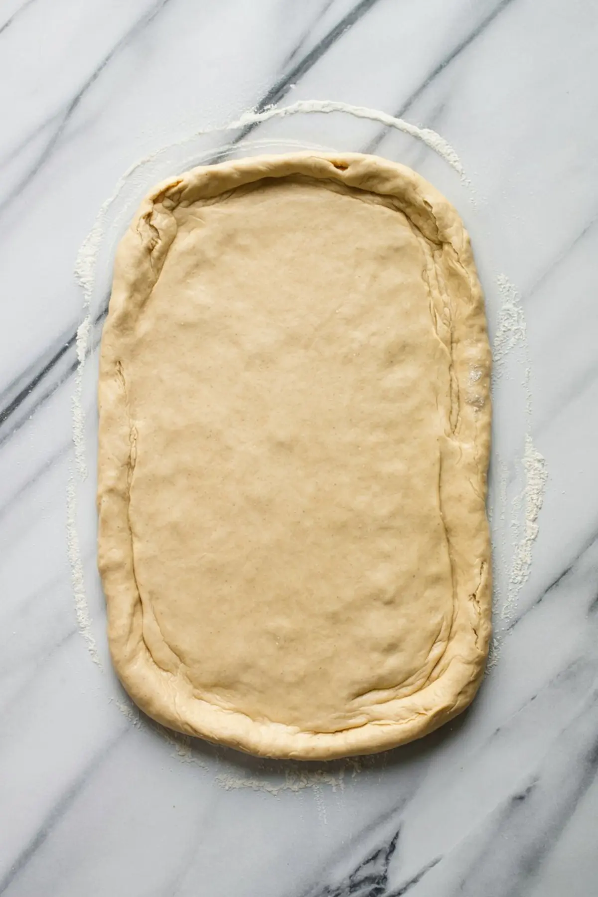 Rectangular sheet of cinnamon roll dough rolled out on a floured marble surface, with slightly raised edges and a smooth, even texture.