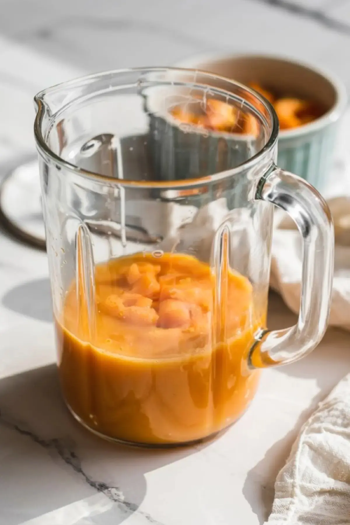 Glass blender pitcher filled with partially blended cantaloupe chunks and juice, placed on a marble surface in natural light.