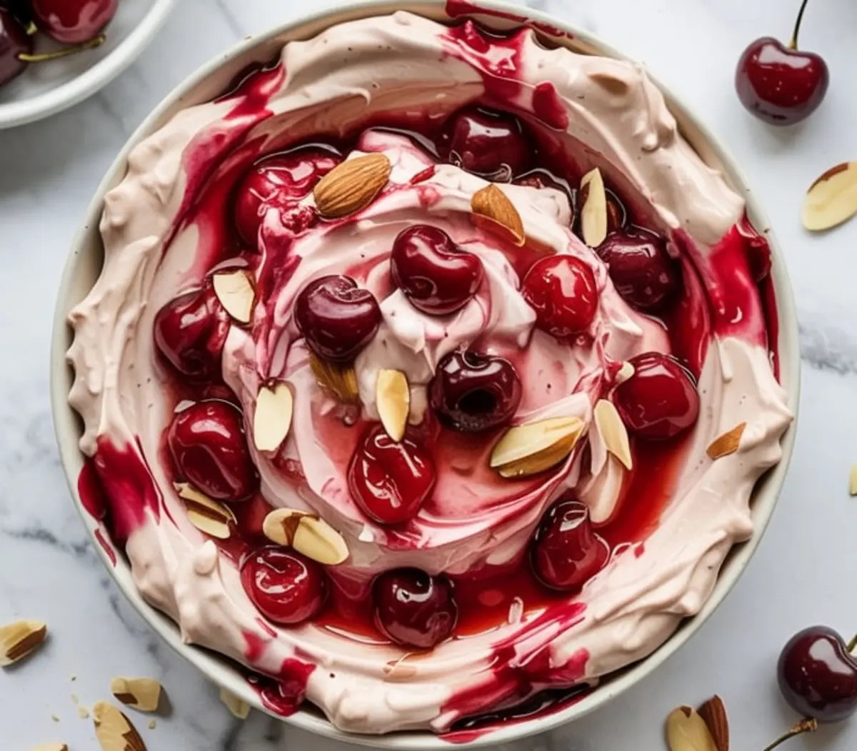 Swirled cherry almond dip in a bowl topped with whole cherries, cherry syrup, and sliced almonds on a marble surface.