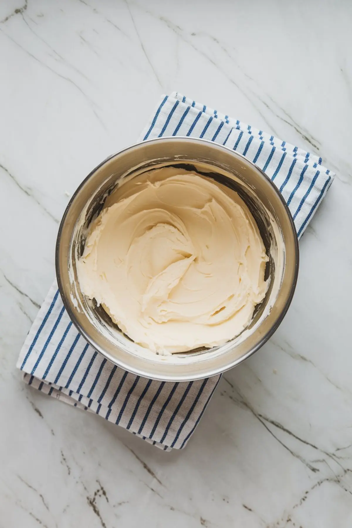 Cream cheese mixture in a metal mixing bowl placed on a blue-striped cloth over a marble surface.