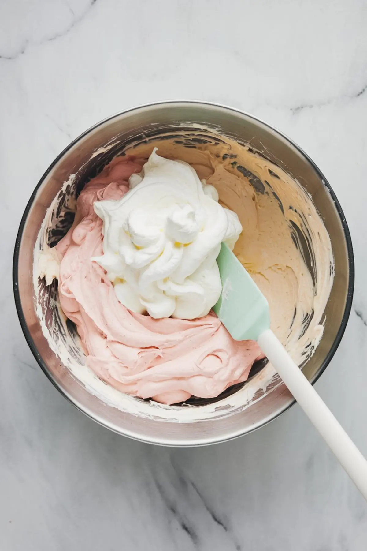 Cherry almond dip mixture in progress inside a metal bowl, with pink and white whipped layers being folded together using a mint green spatula.