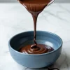 A stream of glossy melted chocolate is being poured from a metal saucepan into a matte blue ceramic bowl on a white marble countertop.