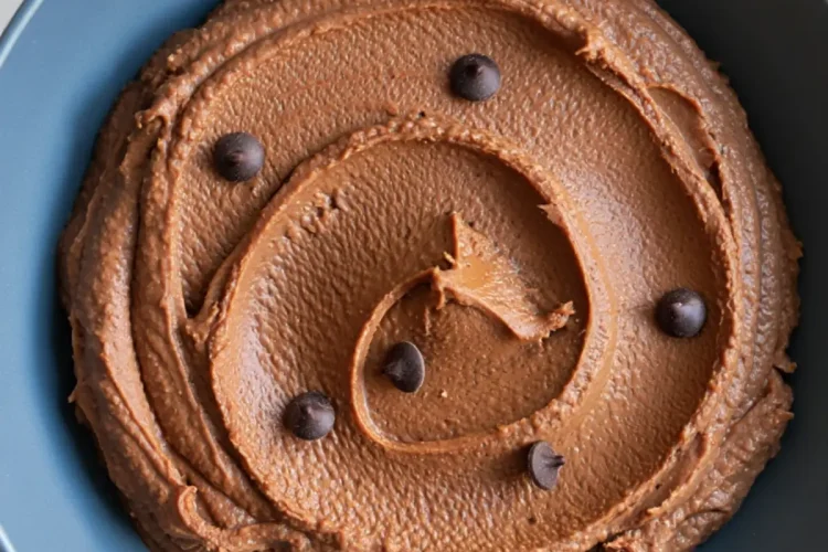 Smooth chocolate hummus swirled in a blue bowl, topped with scattered chocolate chips on a white marble background.