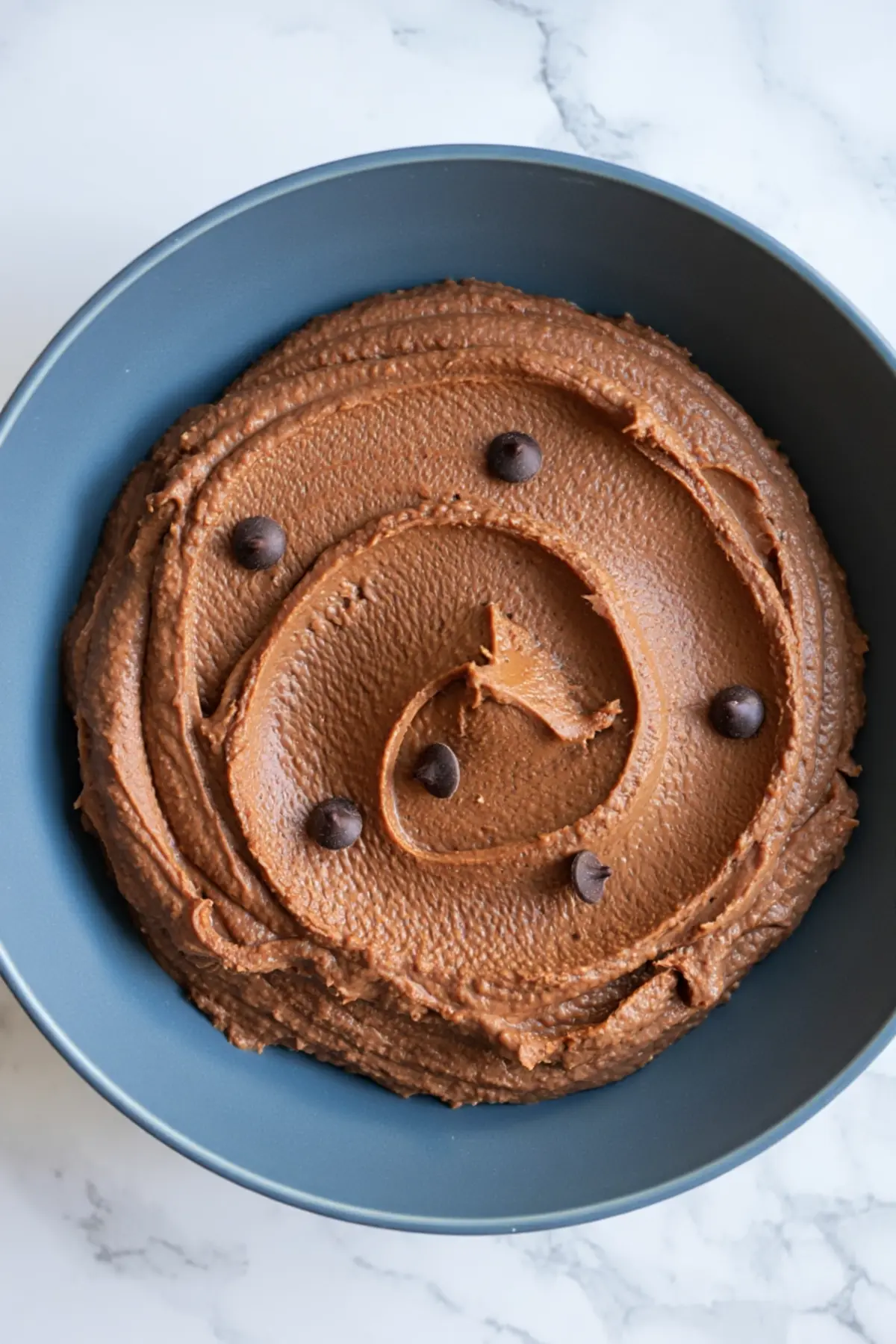 Smooth chocolate hummus swirled in a blue bowl, topped with scattered chocolate chips on a white marble background.