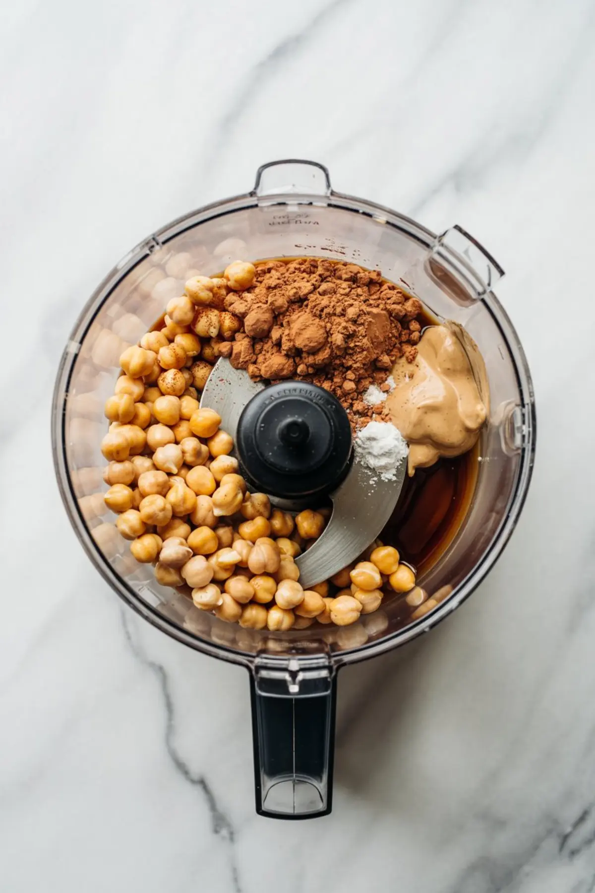 Food processor filled with chickpeas, cocoa powder, peanut butter, maple syrup, and vanilla extract ready to be blended for chocolate hummus.