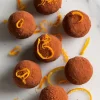 Cocoa-dusted bliss balls topped with orange zest curls, arranged on white marble with scattered citrus peel for a vibrant presentation.