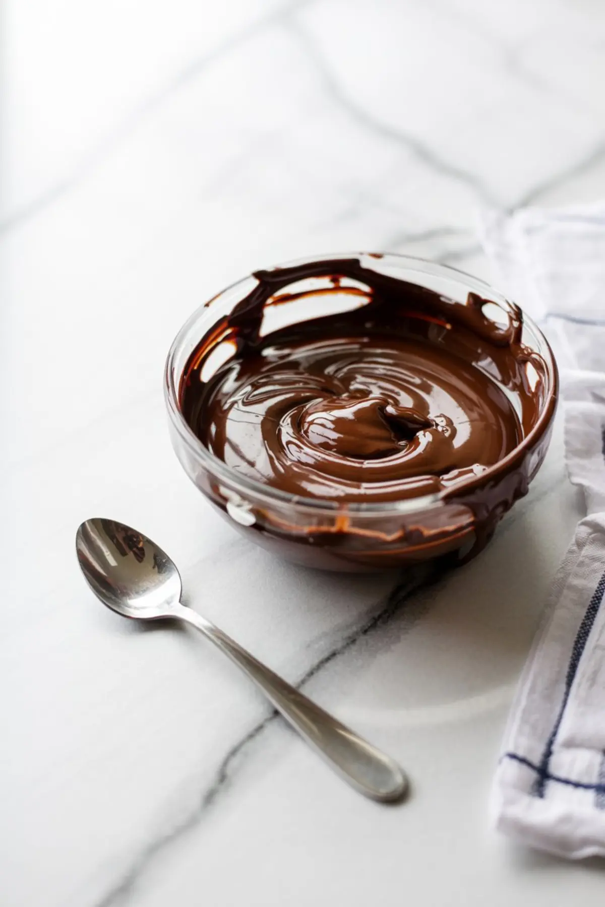 Glass bowl filled with melted chocolate swirled to a glossy finish, set on a white marble surface with a silver spoon and striped kitchen towel.