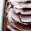 Close-up side angle of a chocolate sheet cake in a white baking dish. The dark chocolate frosting is spread in bold swirls over the dense, moist cake surface.