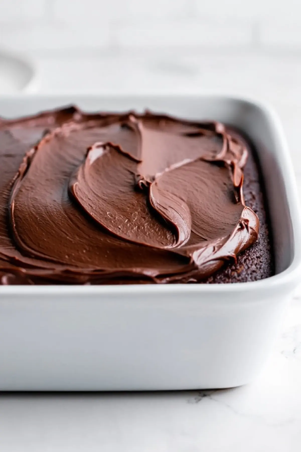 Angled view of a chocolate sheet cake topped with thick, creamy frosting in a smooth swirl pattern. The cake is set in a white rectangular pan on a marble counter.
