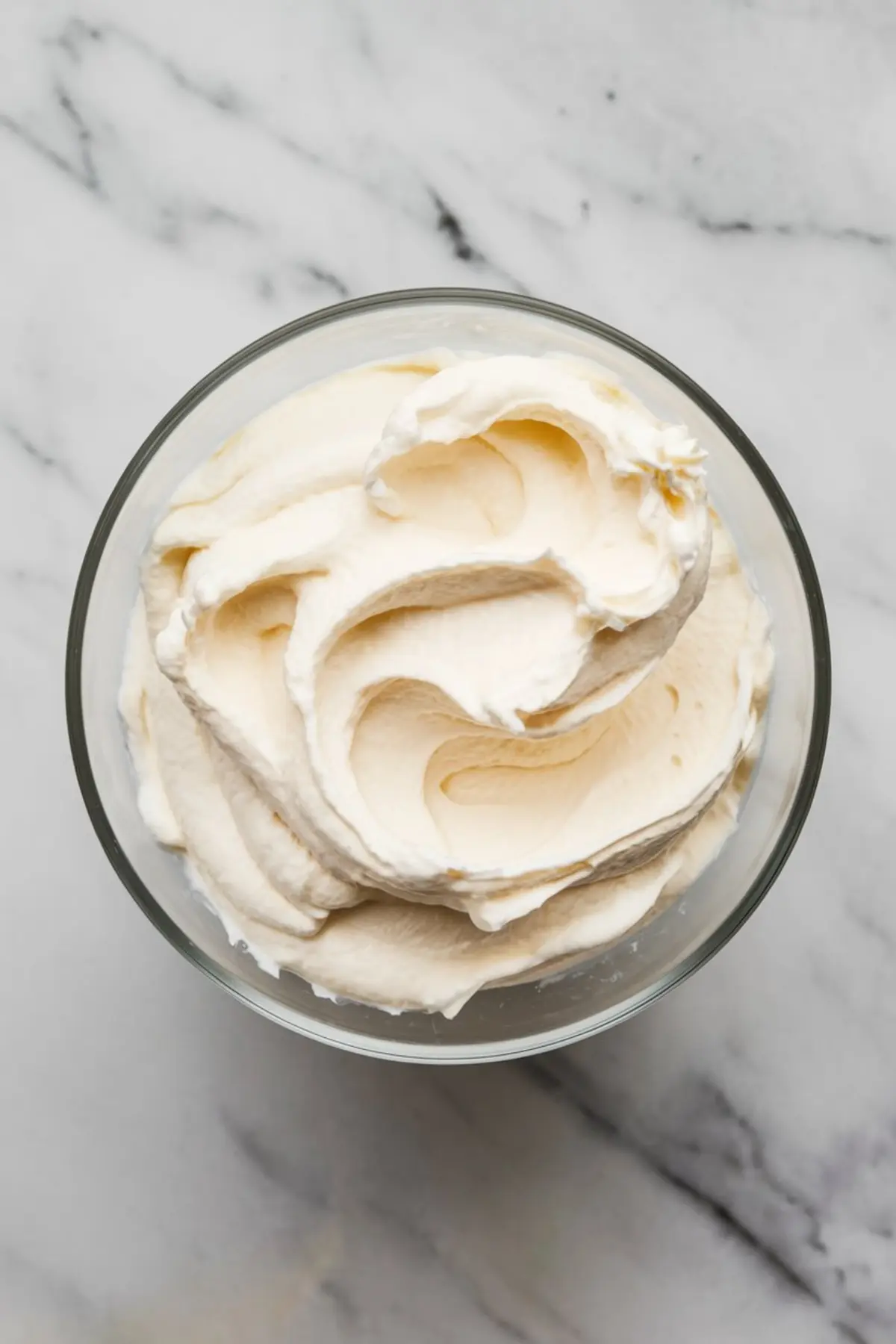 Overhead view of whipped cream in a glass bowl, featuring soft, smooth swirls with a creamy texture, ideal for desserts or toppings on cakes and puddings.