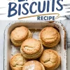 Golden brown biscuits stacked in a rustic white enamel baking tray, showcasing a fluffy, layered texture with a cracked top crust and a dusting of flour, set against a marble countertop.