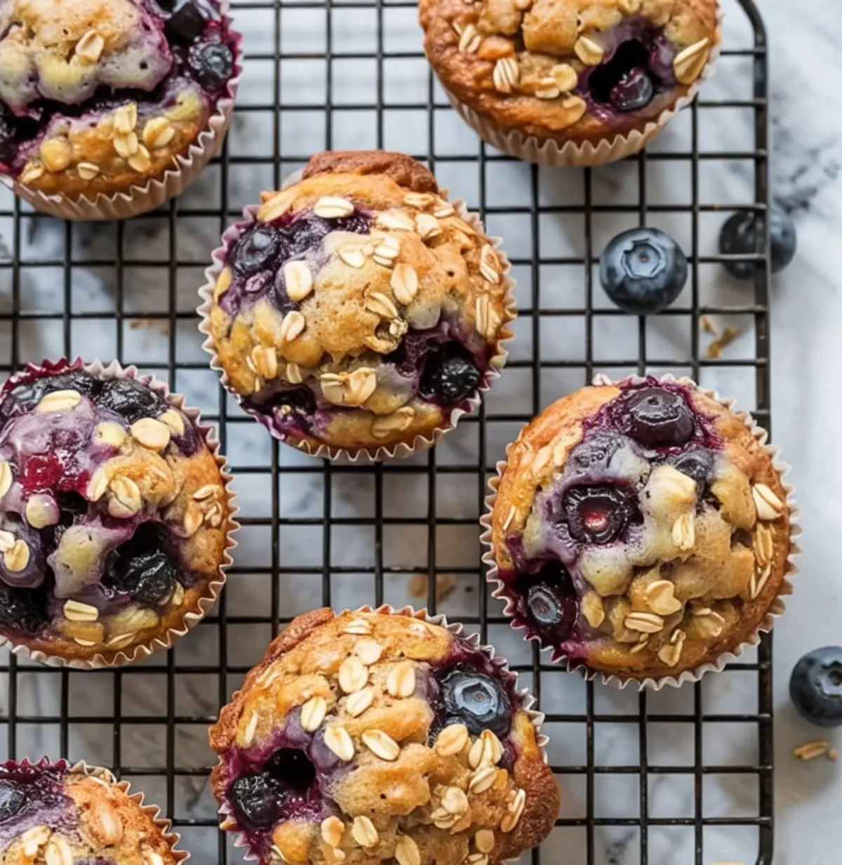 Close-up top view of blueberry oat muffins with golden tops and oat flakes, cooling on a black wire rack over parchment paper, surrounded by scattered blueberries.