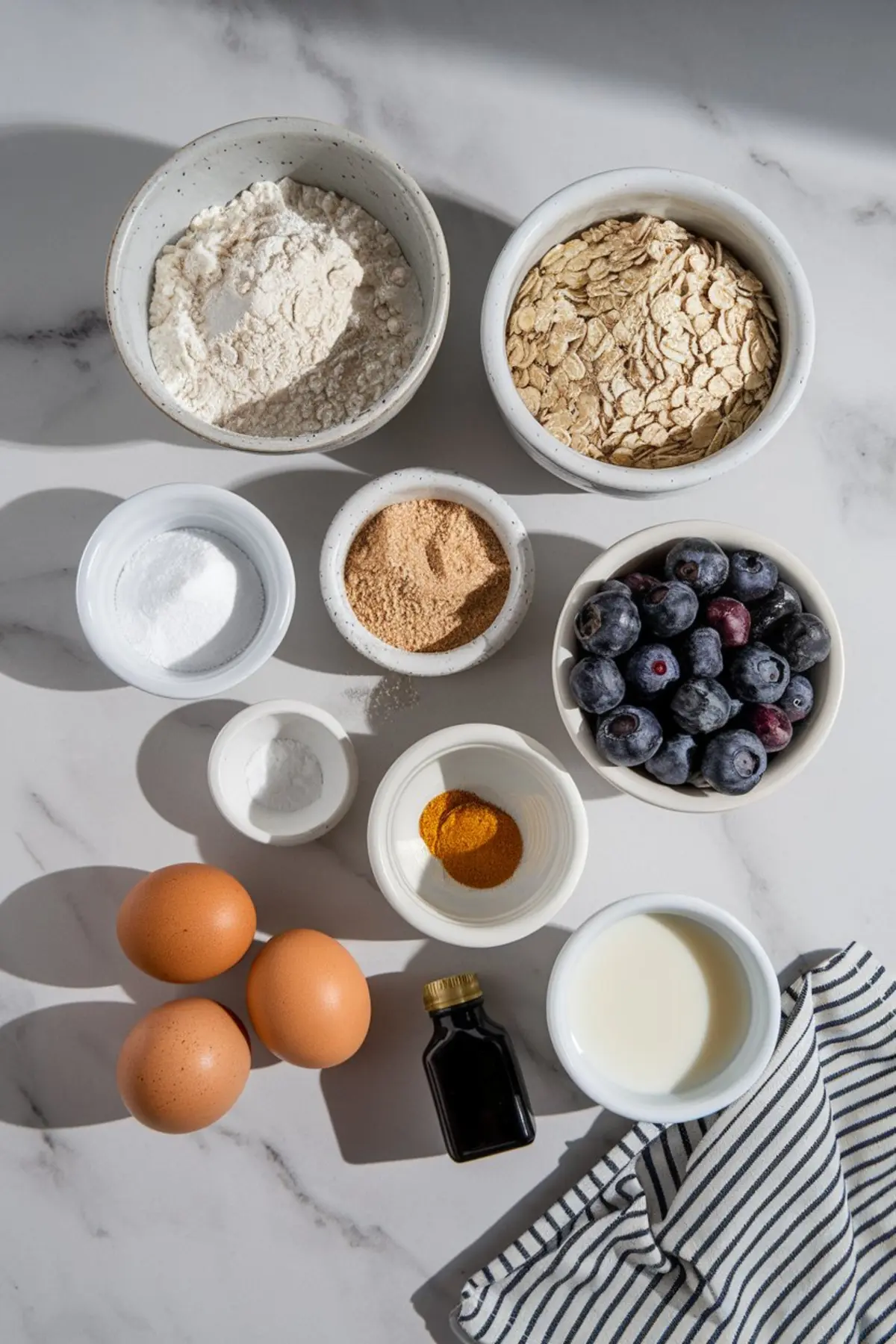 Flat lay of blueberry oat muffin ingredients on a marble surface, including flour, oats, fresh blueberries, brown sugar, baking powder, turmeric, eggs, milk, vanilla extract, and a striped kitchen towel.