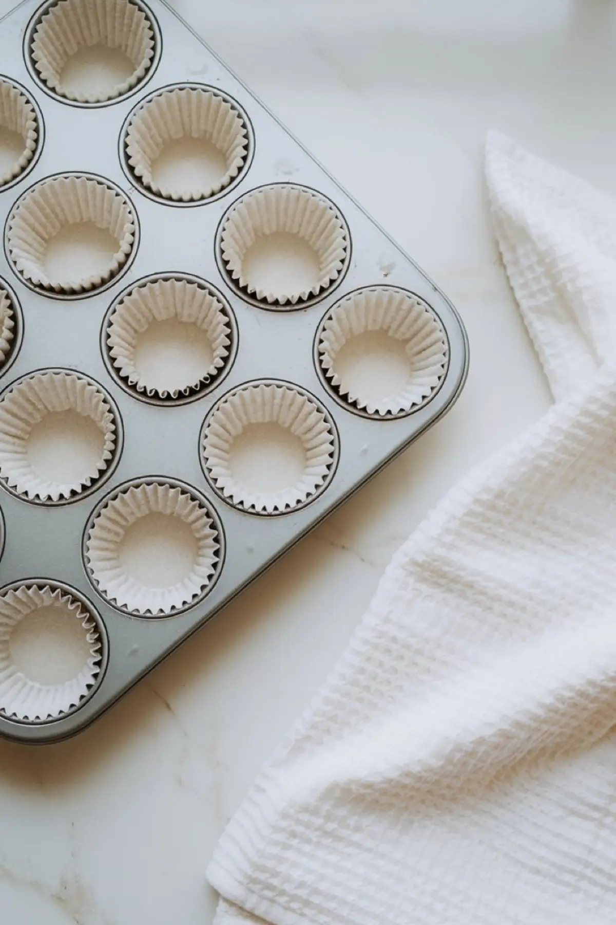 Muffin tin lined with white paper cupcake liners, placed on a marble surface beside a textured white kitchen towel, prepped for baking.