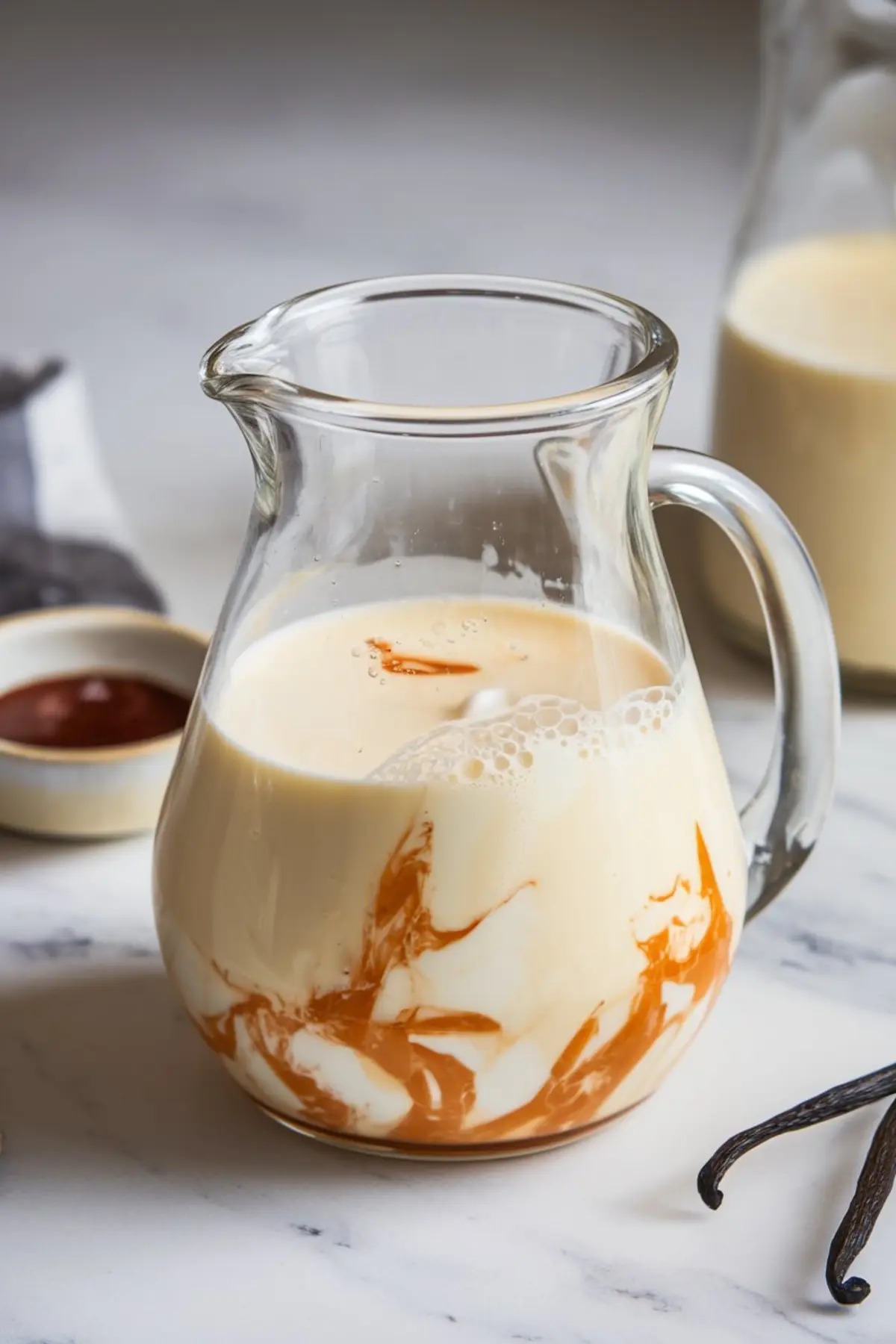 Glass pitcher containing the signature tres leches mixture with visible swirls of caramel and milk, placed on a marble counter near a vanilla bean and small dish.
