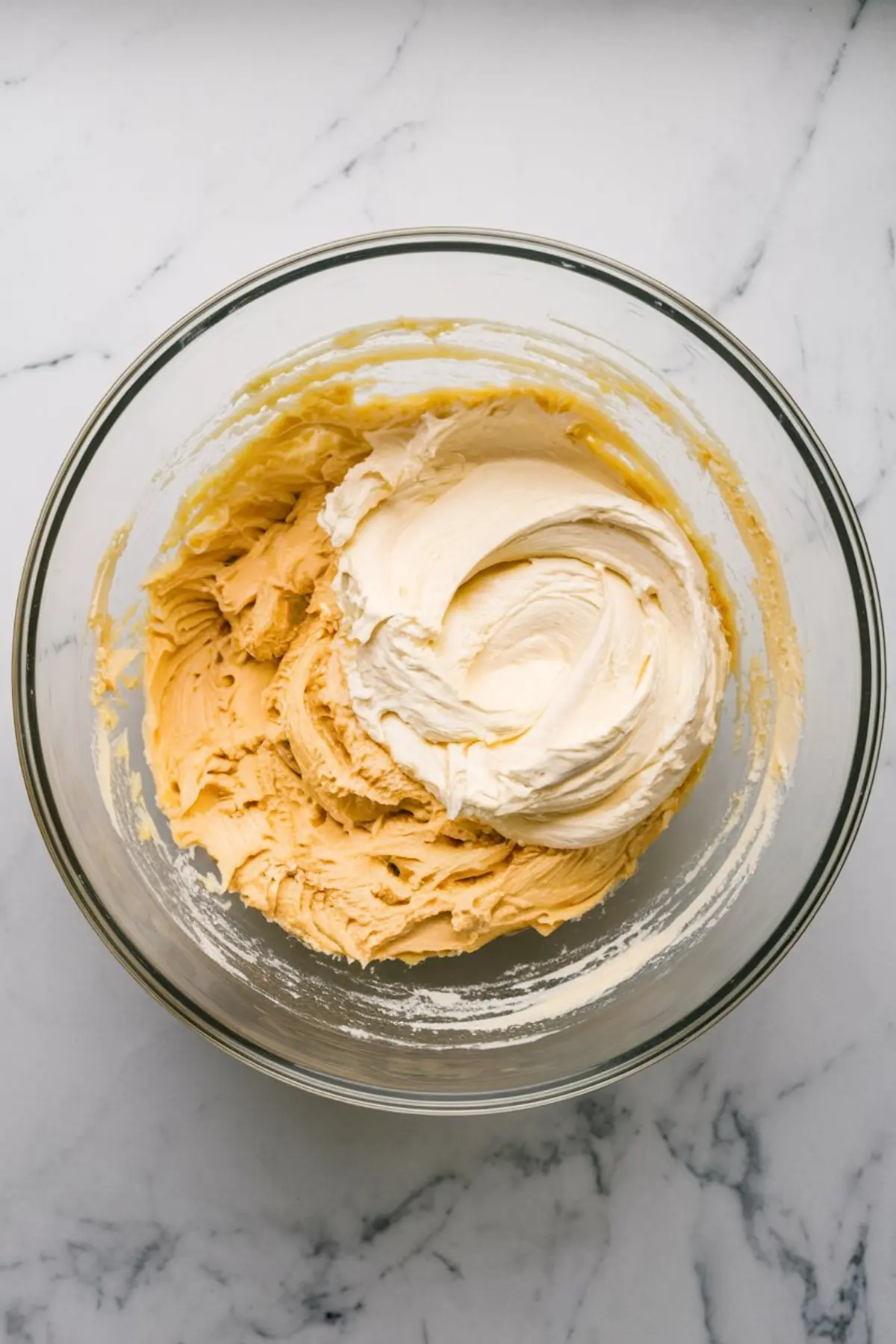 Glass mixing bowl with swirled layers of cheesecake batter and a golden-brown cookie dough mixture, placed on a marble countertop.
