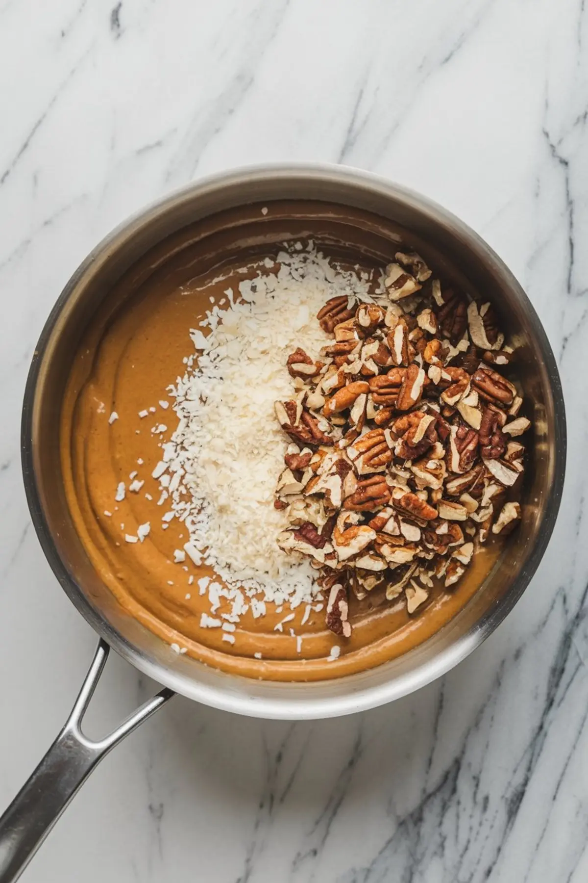 Overhead view of a saucepan filled with thick caramel-colored frosting, topped with shredded coconut and chopped pecans, ready to be mixed for German chocolate cake.