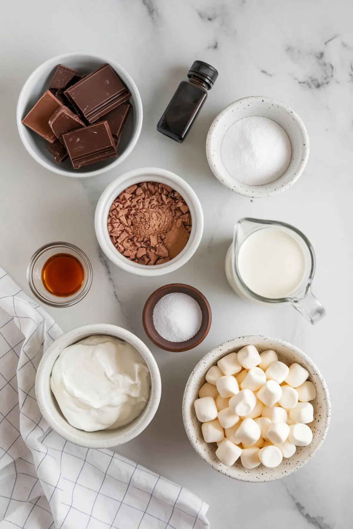 Flat lay of hot chocolate ingredients arranged on a white marble surface. Includes dark chocolate squares, cocoa powder, mini marshmallows, granulated sugar, milk, salt, vanilla extract, and heavy cream in ceramic and glass bowls, styled for a cozy baking or winter drink preparation theme.
