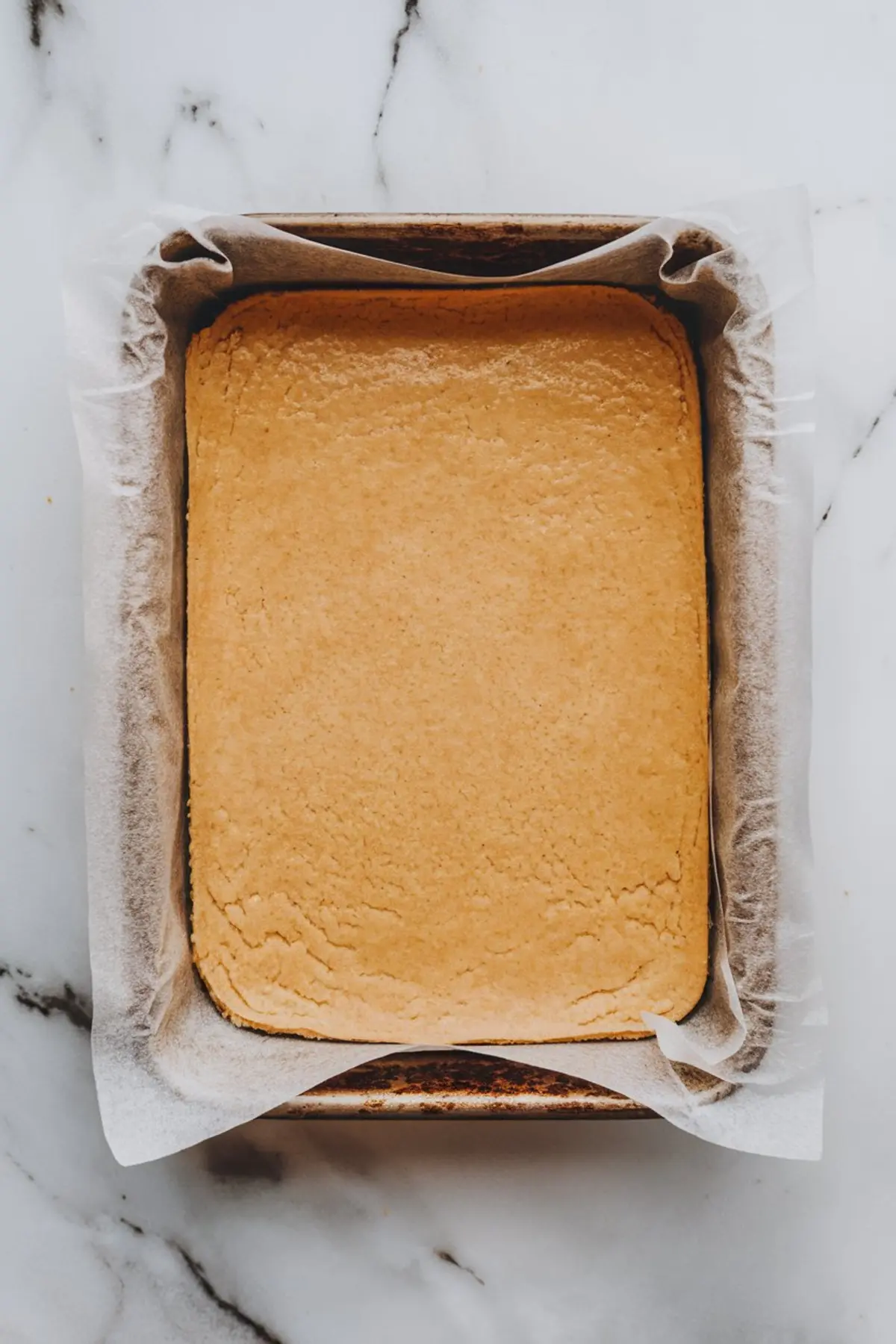 Golden shortbread crust baked in a parchment-lined rectangular baking pan, shown cooling on a marble counter.