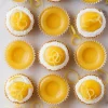 Overhead image of decorated and undecorated lemon cookie cups arranged in rows, with some topped with white icing and lemon zest curls on a white background scattered with lemon peel ribbons.