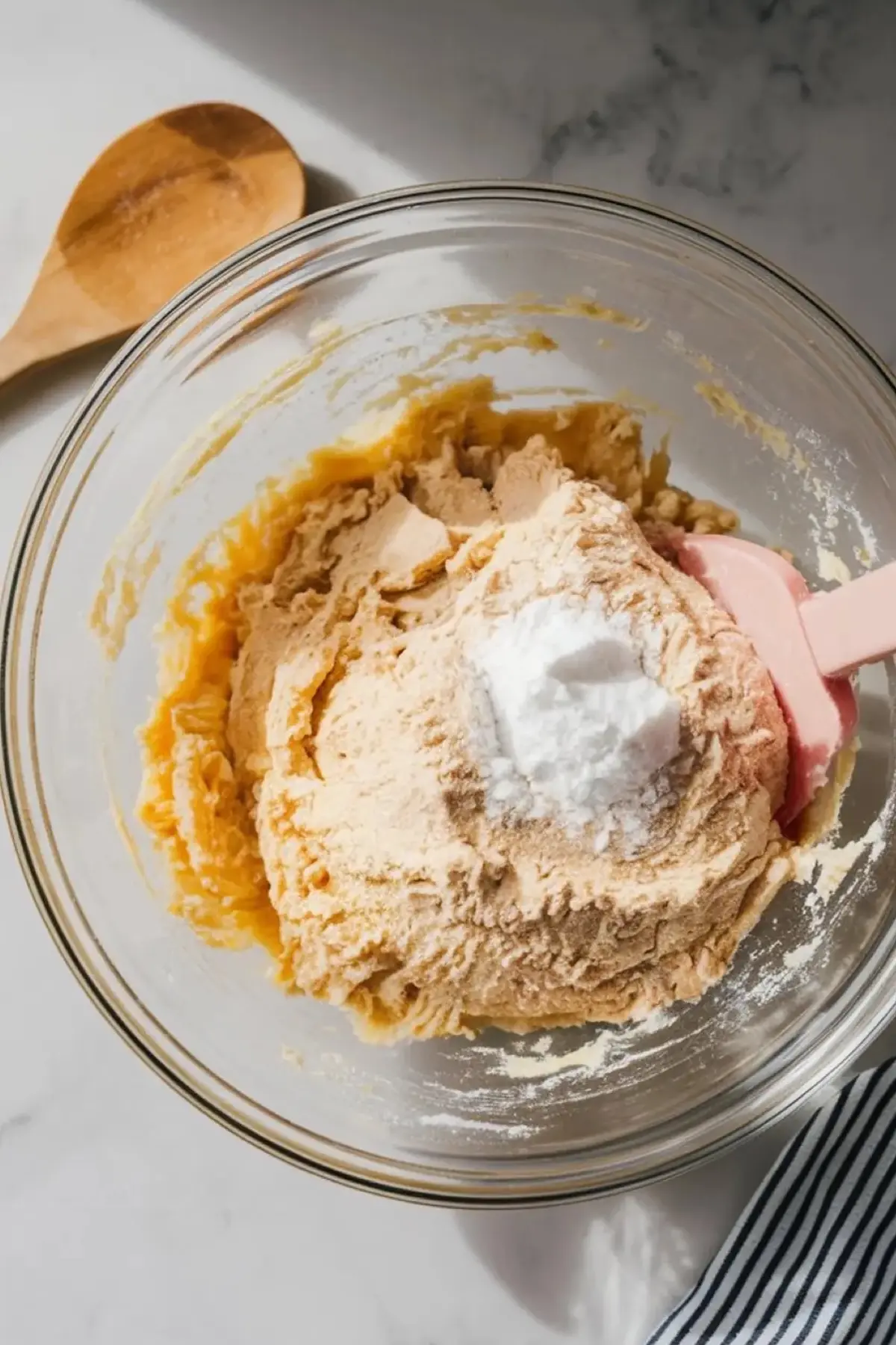 Mixing bowl with cookie dough in progress, topped with a mound of flour and baking powder, and a pink spatula resting in the mixture on a marble countertop.