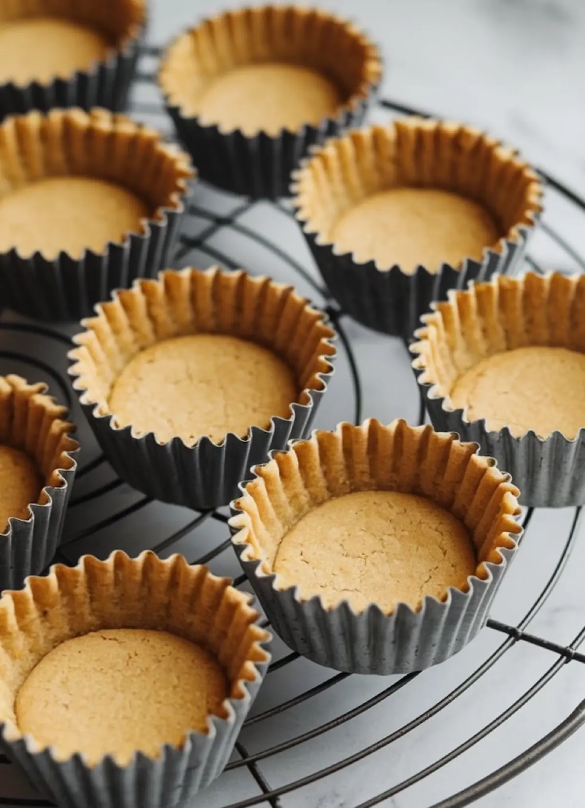 Baked cookie cup shells cooling on a black wire rack, each with a golden-brown center and ridged edges in dark paper liners.