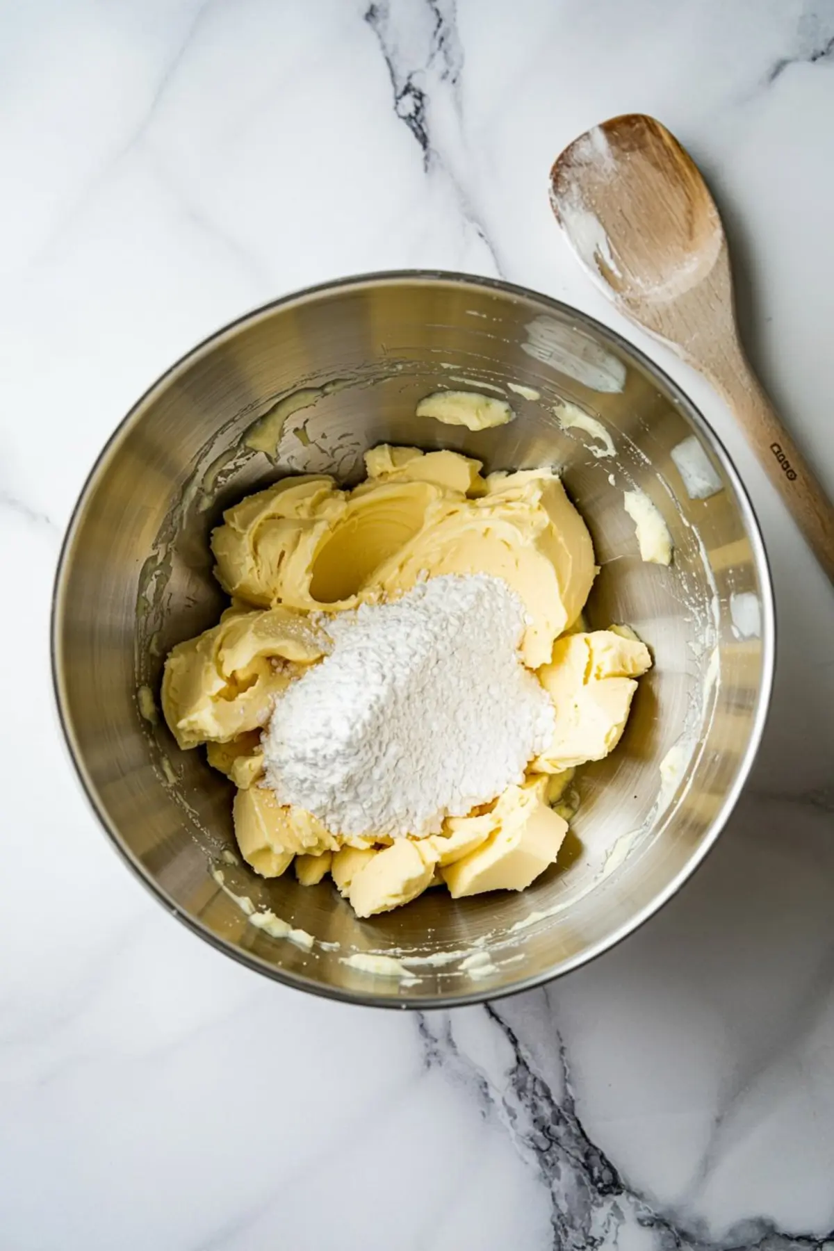 Mixing bowl filled with softened butter and powdered sugar, showing the early creaming stage of sugar cookie dough preparation with a wooden spoon nearby.