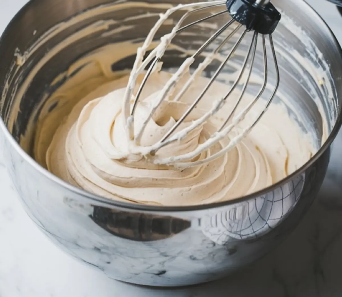 Whipped frosting in a stainless steel mixing bowl with a whisk attachment coated in fluffy lemon frosting, resting on a white countertop.