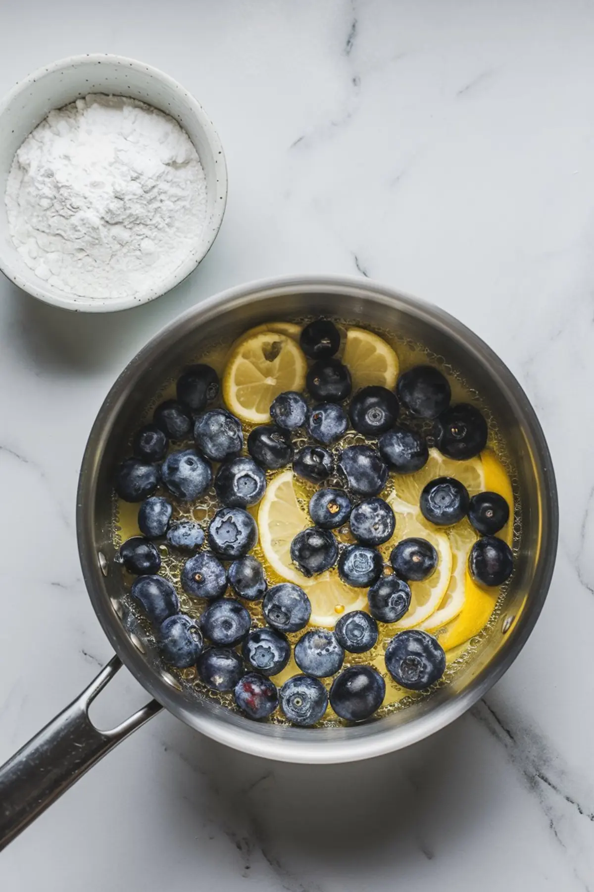 Fresh blueberries and lemon slices simmering in a saucepan with sugar and liquid, next to a bowl of cornstarch, illustrating the cooking of homemade blueberry lemon compote.