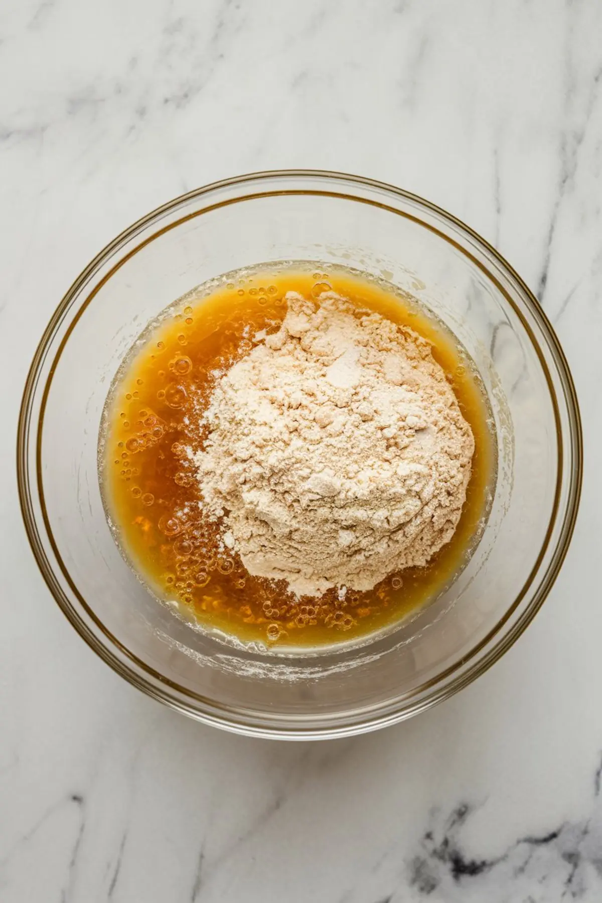Glass bowl containing wet ingredients including oil, eggs, and vanilla, partially covered with dry ingredients ready for mixing, captured from a top-down view on marble.