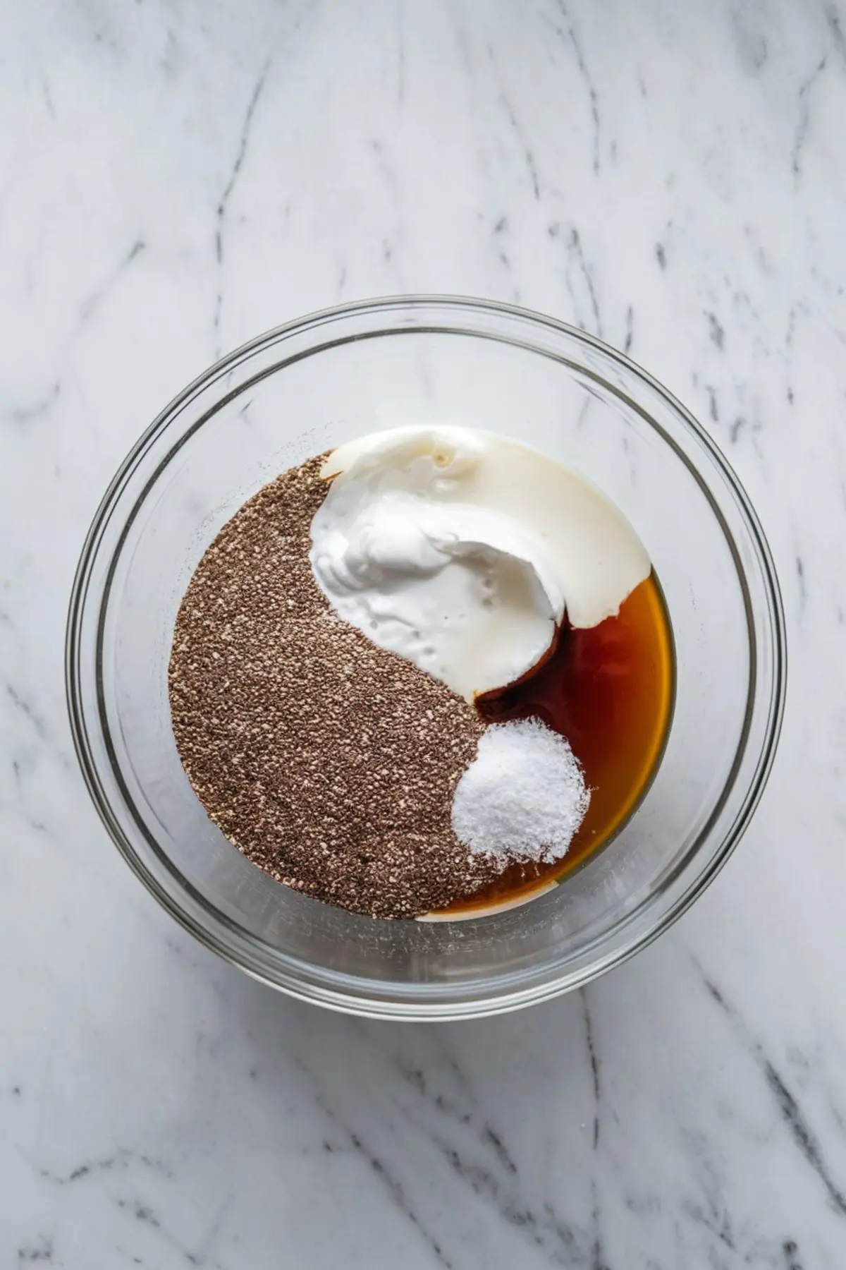 Unmixed ingredients for mango chia pudding in a glass bowl, including chia seeds, yogurt, maple syrup, and a mound of salt on a marble background.