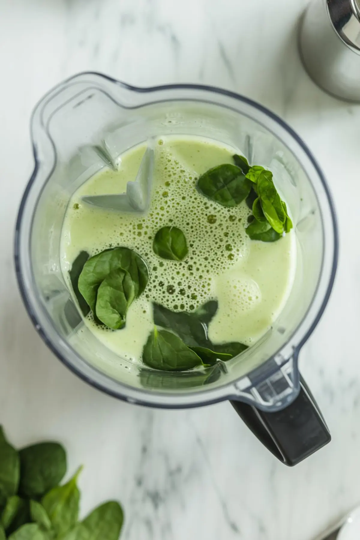 Overhead view of a blender with a green smoothie mixture and fresh spinach leaves visible on top, showing the preparation of the green Tajín layer for layered mango popsicles.