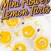 Decorative mini flower-shaped lemon tarts with bright yellow filling and powdered sugar edges, garnished with lemon slices and edible flowers on a white platter.