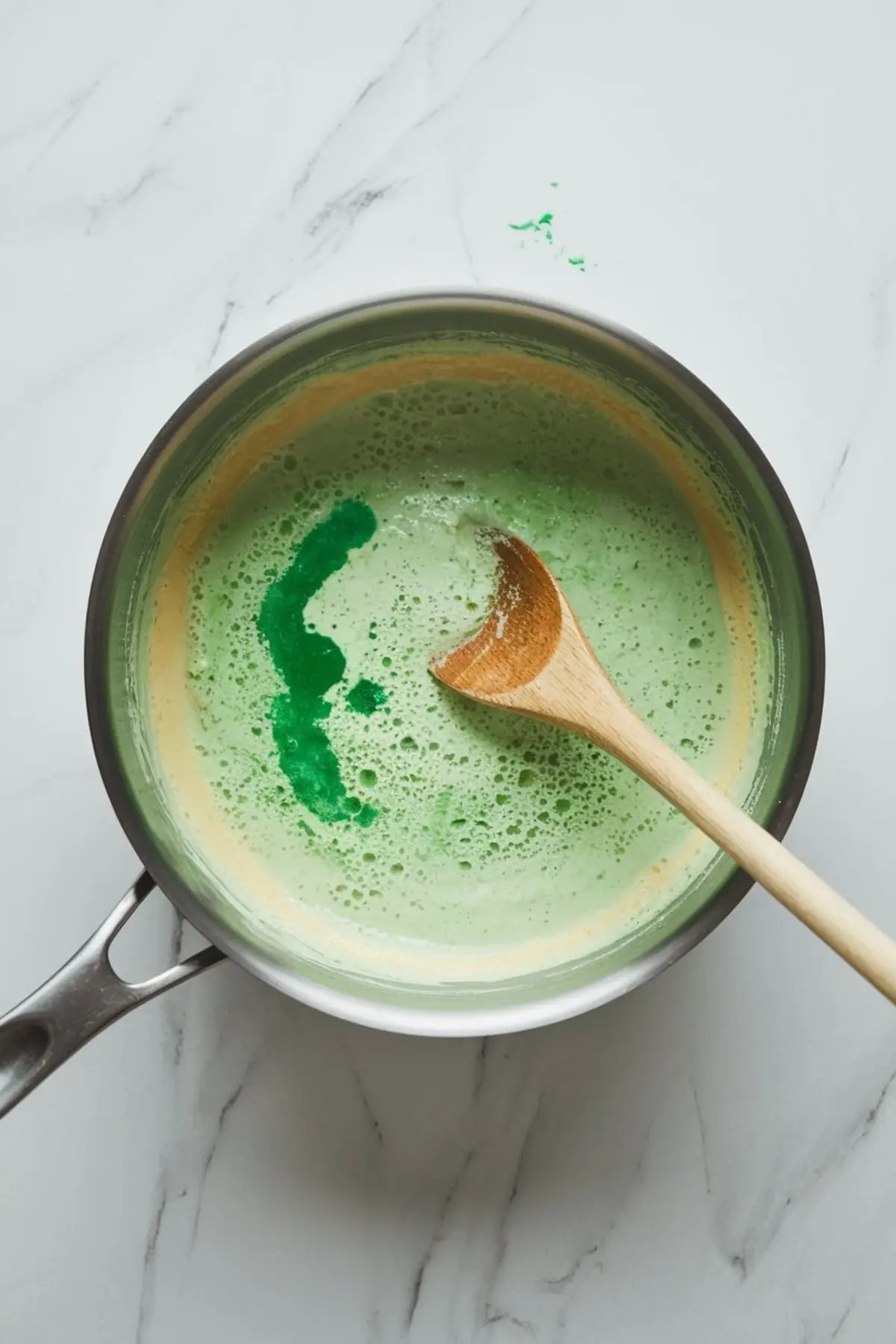 Stainless steel pot containing green-tinted custard mixture swirled with food coloring and a wooden spoon, captured on a clean white marble background during mint flavor infusion.
