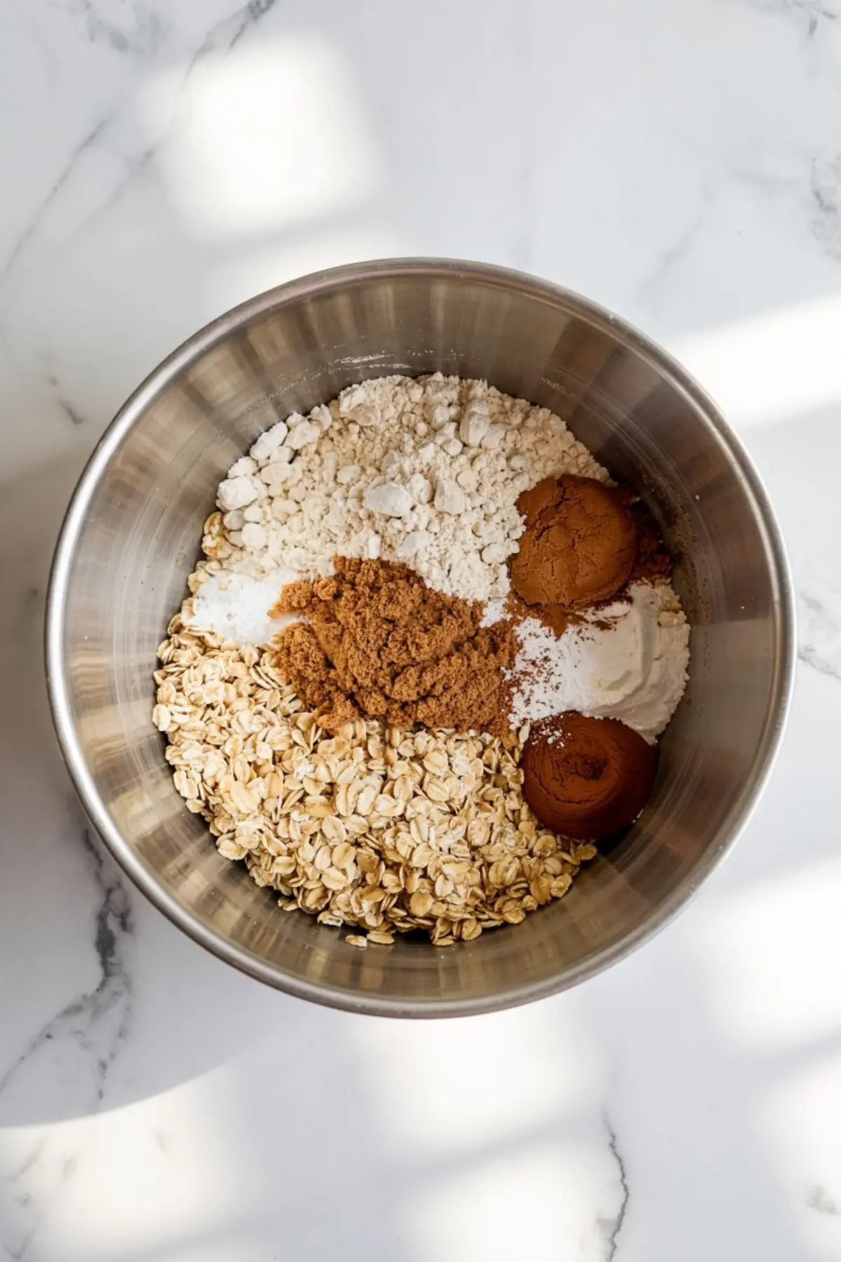 Stainless steel mixing bowl filled with dry ingredients including rolled oats, brown sugar, cinnamon, flour, baking powder, and salt, set on a white marble surface.
