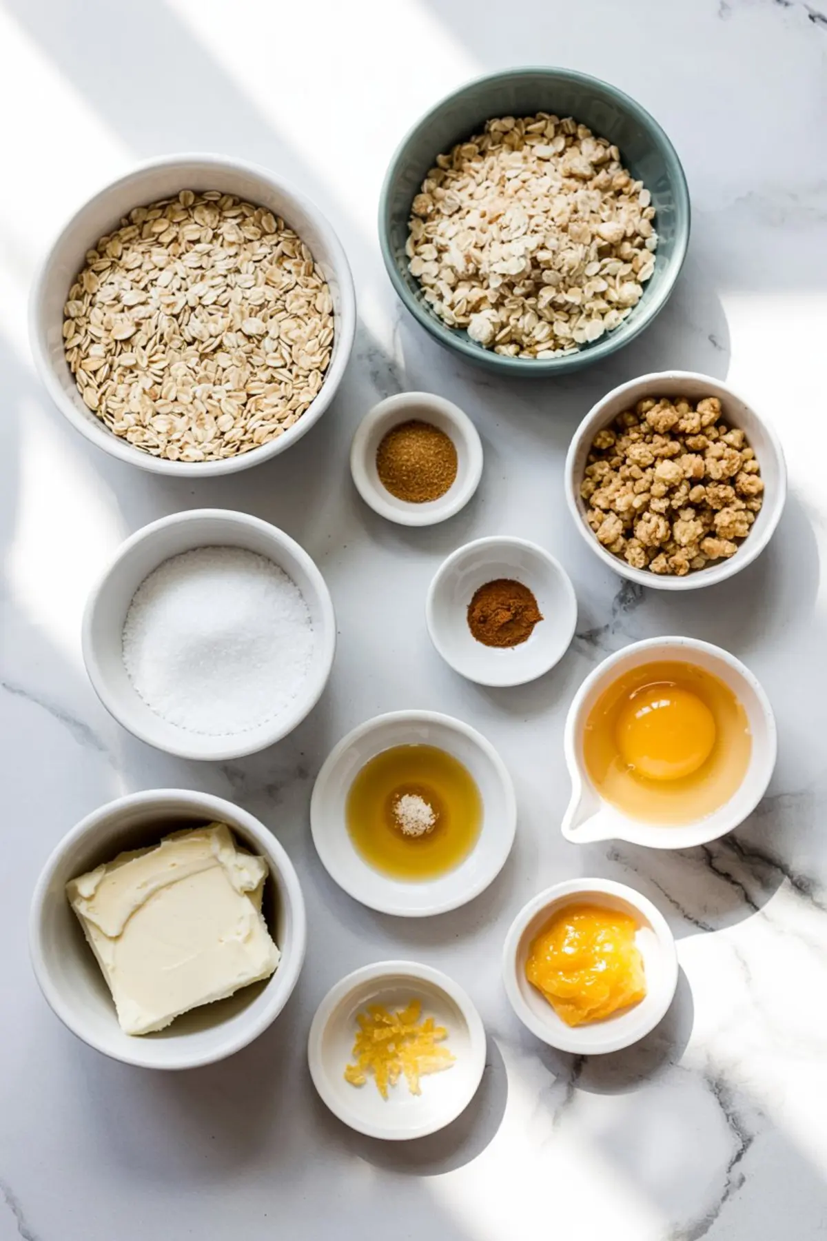 Overhead view of individual baking ingredients for oatmeal lemon bars arranged in small bowls, including oats, cream cheese, lemon curd, egg yolk, vanilla, cinnamon, and brown sugar.