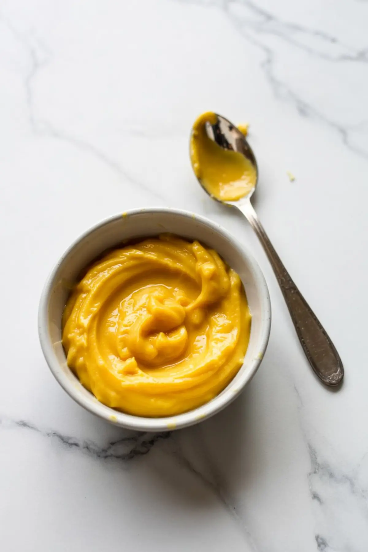 Small white bowl filled with smooth, glossy lemon curd and a spoon resting nearby with remnants of the bright yellow spread on a marble countertop.