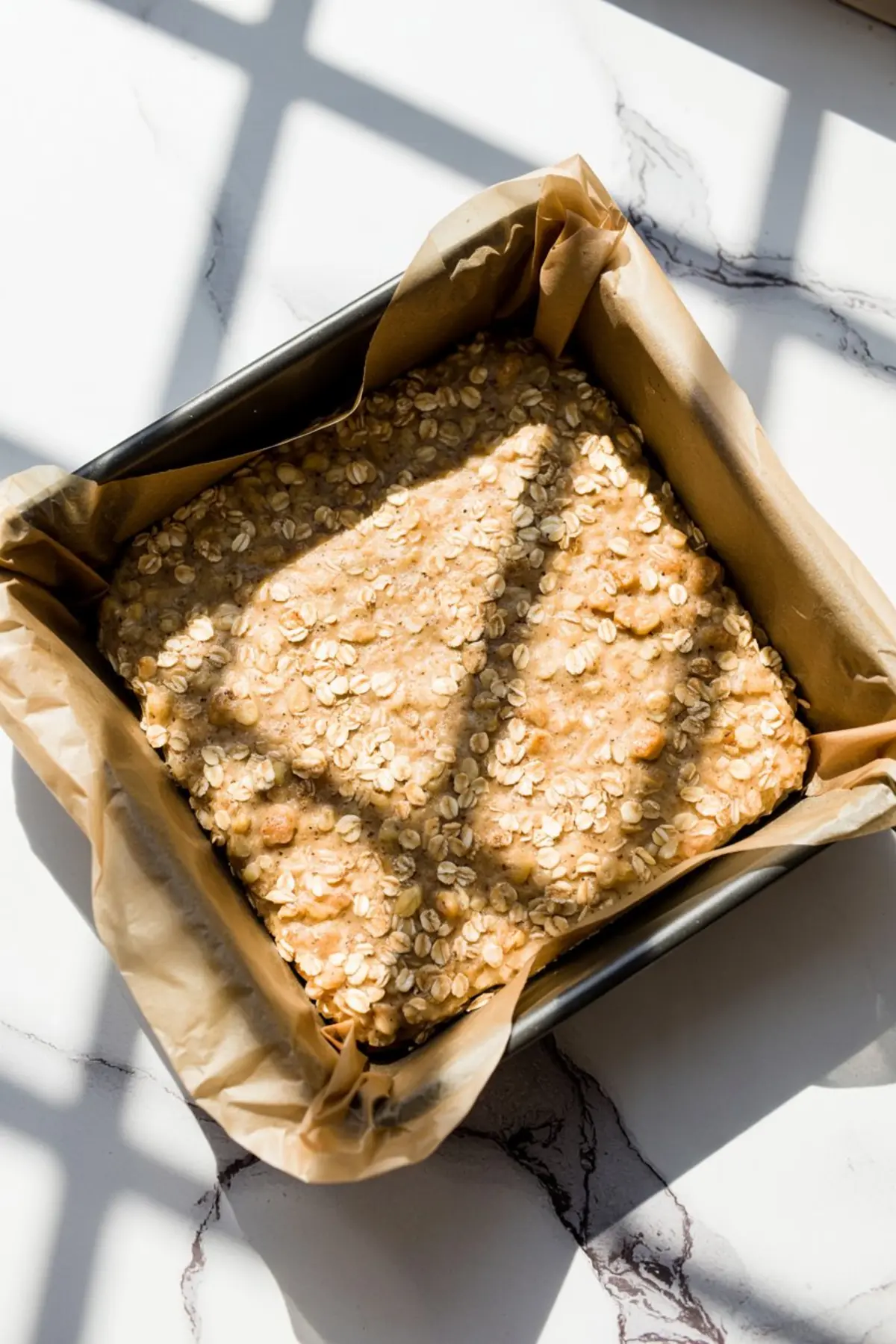 Square baking pan lined with parchment paper and filled with unbaked oatmeal crumble mixture, showing an even layer of oat-studded dough pressed into the bottom.