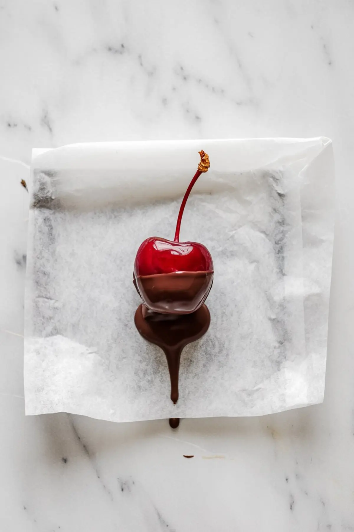 Single cherry dipped halfway in melted chocolate, placed on a parchment-lined surface with chocolate dripping down.
