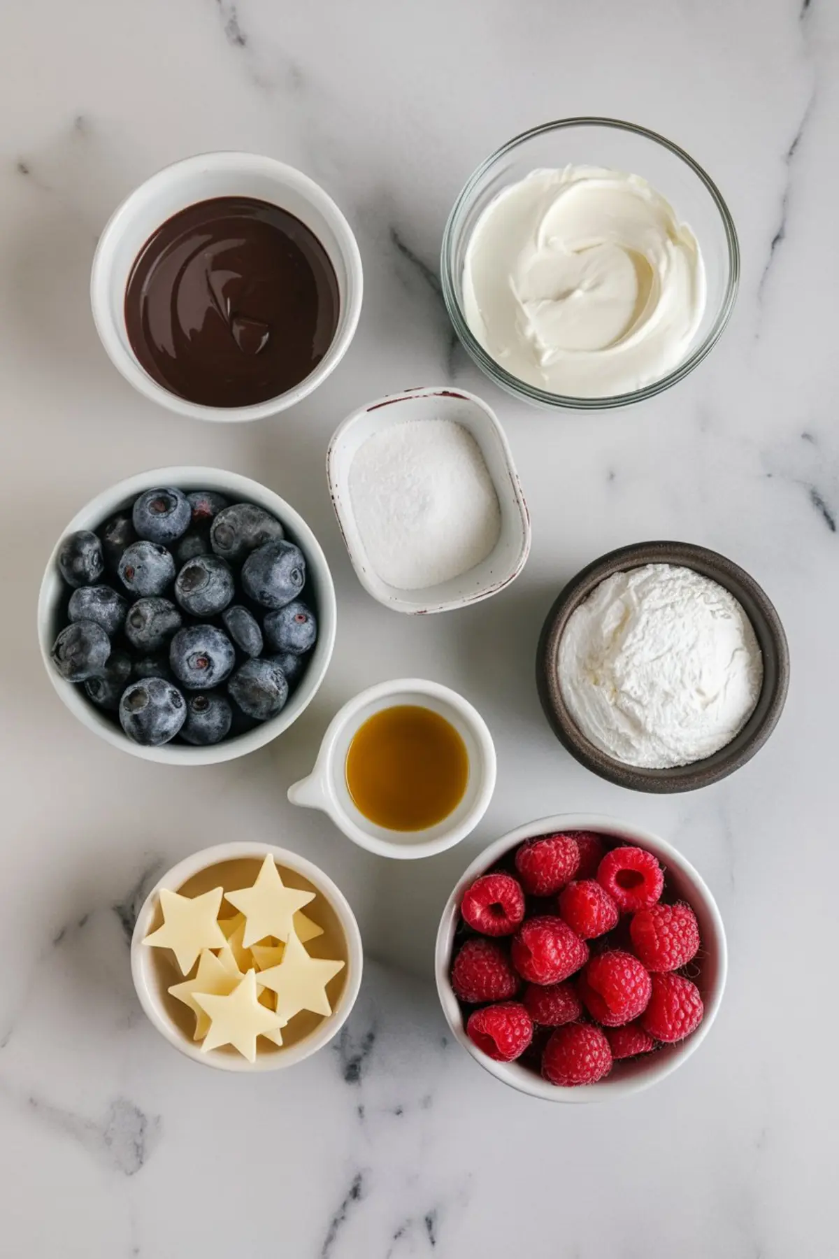 Flat lay of tartlet ingredients including fresh blueberries, raspberries, melted chocolate, powdered sugar, cream cheese, vanilla extract, white chocolate stars, and heavy cream on a white marble background.