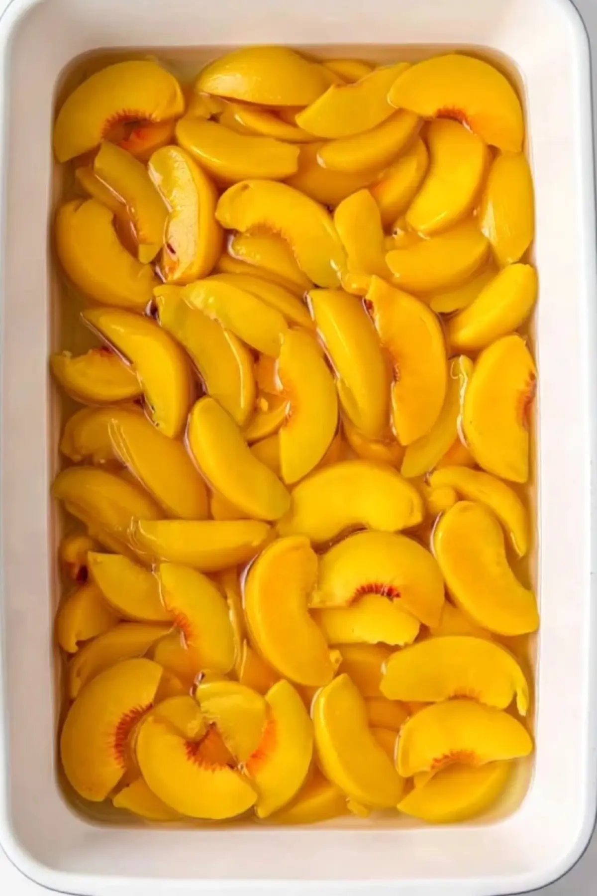 White baking dish filled with sliced canned peaches in syrup, arranged in a single layer, ready to be topped and baked for a peach cobbler dessert.