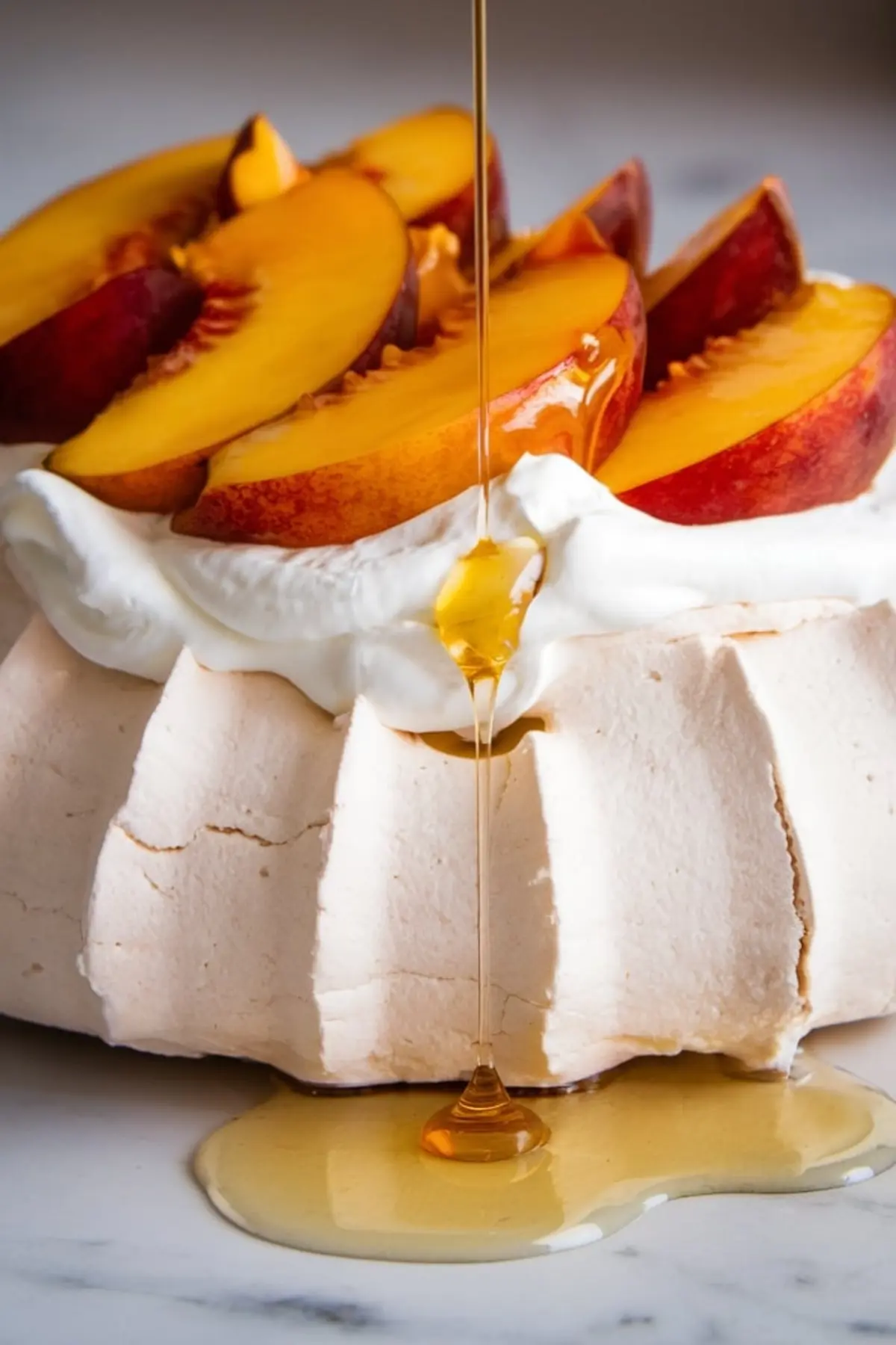 Close-up of honey pouring onto peach pavlova with a swirl of whipped cream. Fresh peach wedges add a vibrant, fruity garnish to the crisp meringue shell.