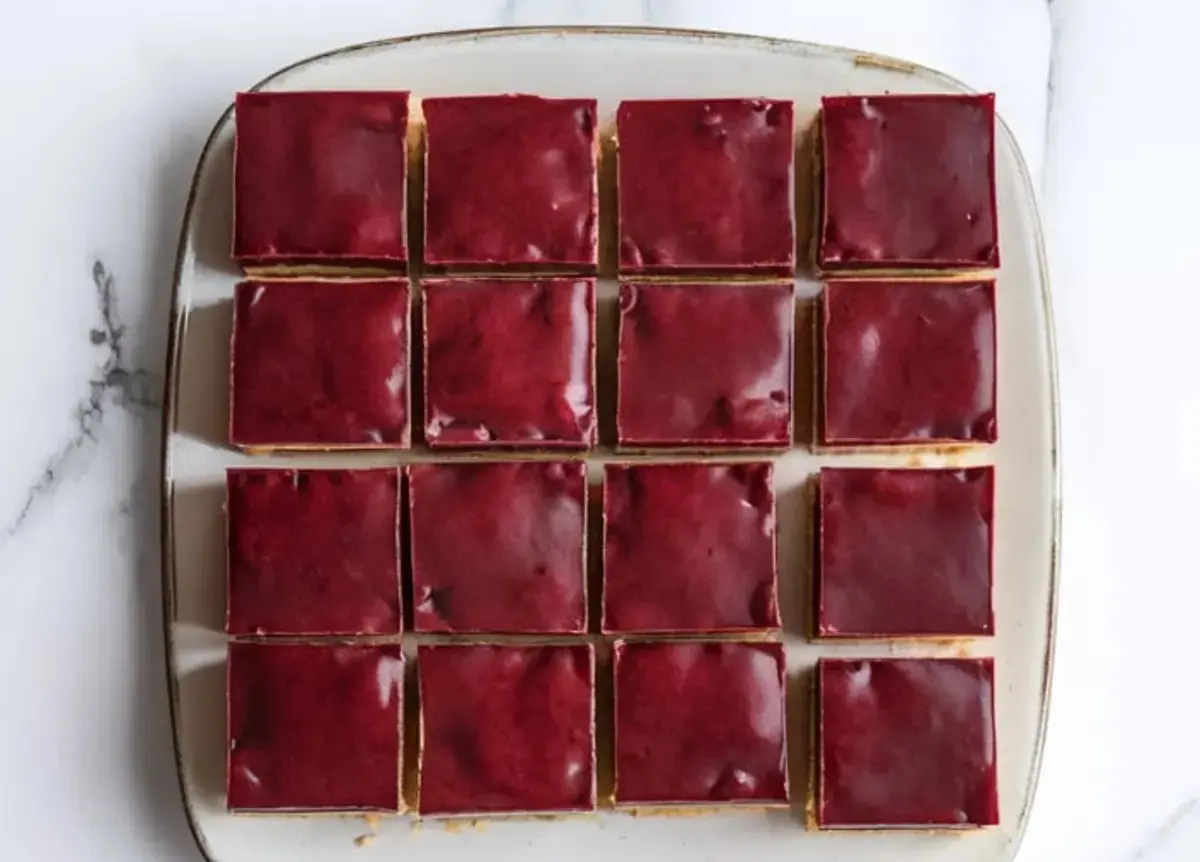 Overhead view of neatly arranged raspberry caramel millionaire’s shortbread squares on a white plate, each bar featuring a glossy raspberry chocolate top layer, creamy caramel center, and a golden shortbread base.