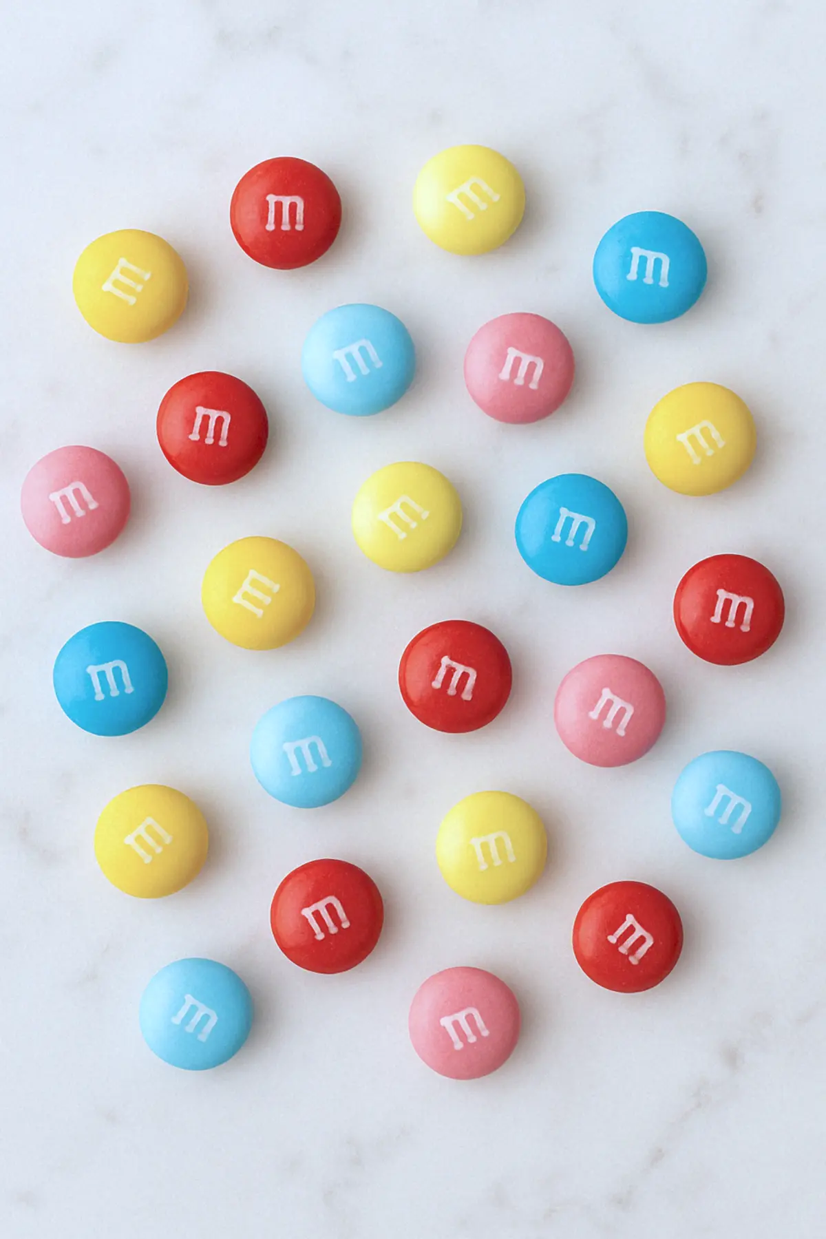 Flat lay of pastel-colored M&M candies in red, yellow, pink, and blue, scattered across a white marble surface, ideal for decorating desserts and adding color to sweet treats.

