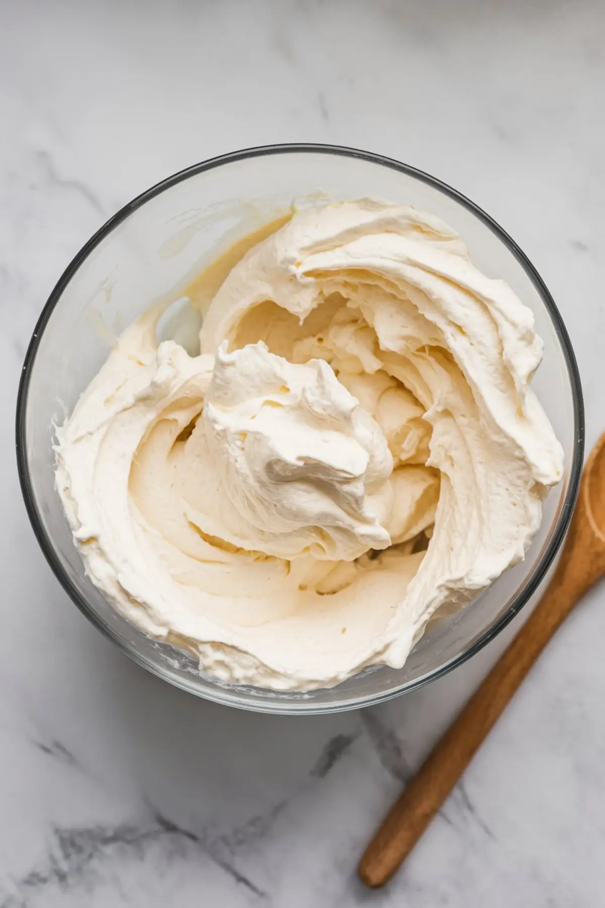 Bowl of whipped topping with a smooth and airy texture, shown with a wooden spoon nearby, set on a light marble surface, ideal for dessert dip recipes and frostings.