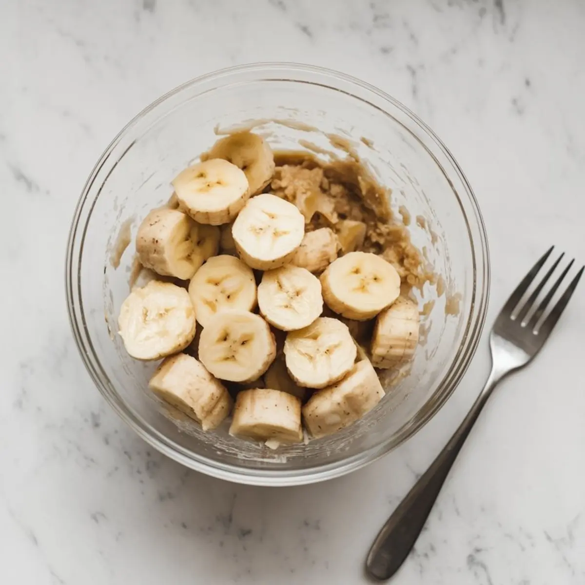 Sliced ripe bananas in a glass bowl on a white marble surface, ready to be mashed as a base for banana muffin batter.