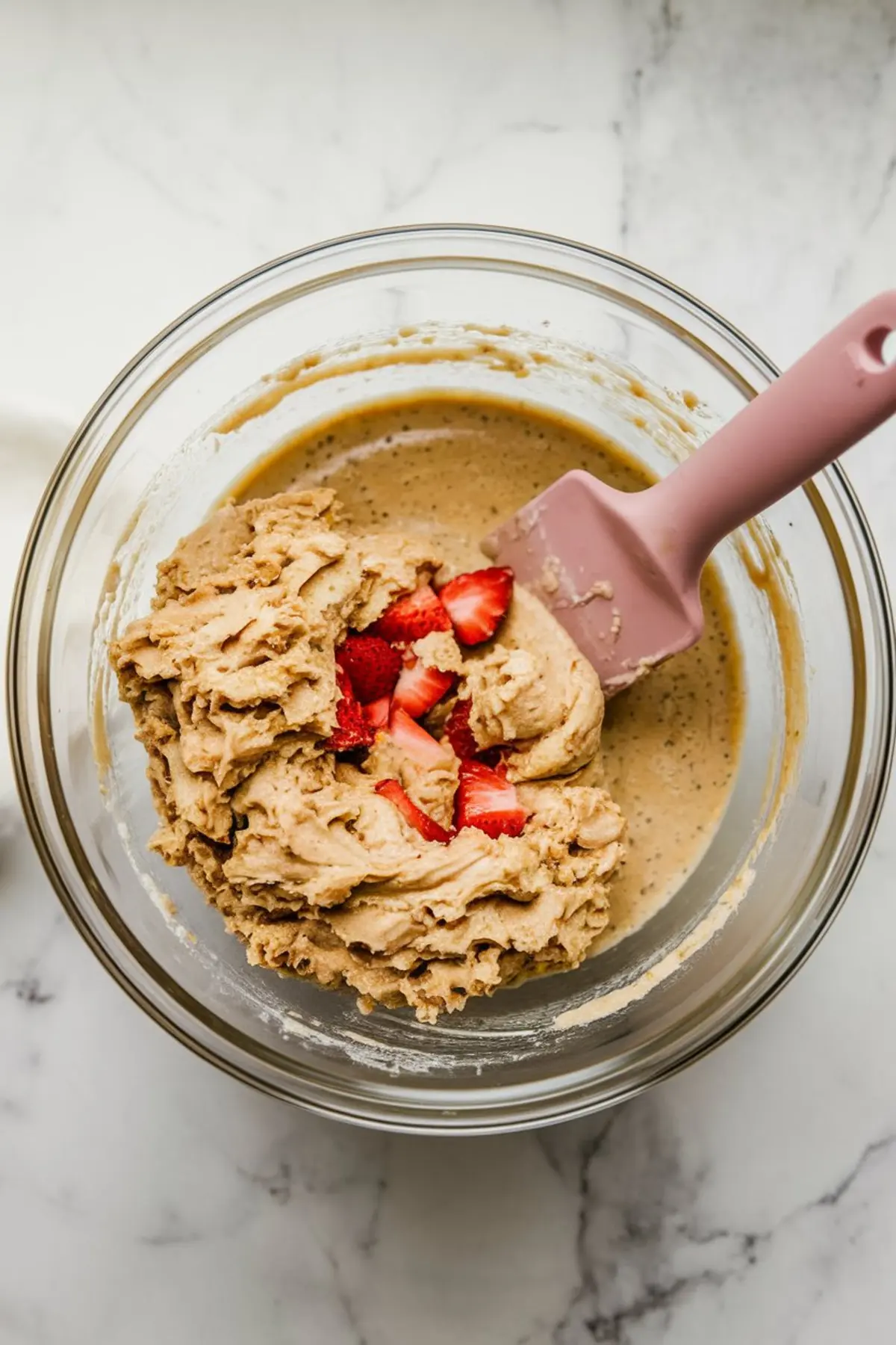 Partially mixed muffin batter in a glass bowl with chopped strawberries on top and a pink spatula resting inside, ready to be folded in for strawberry banana muffins.