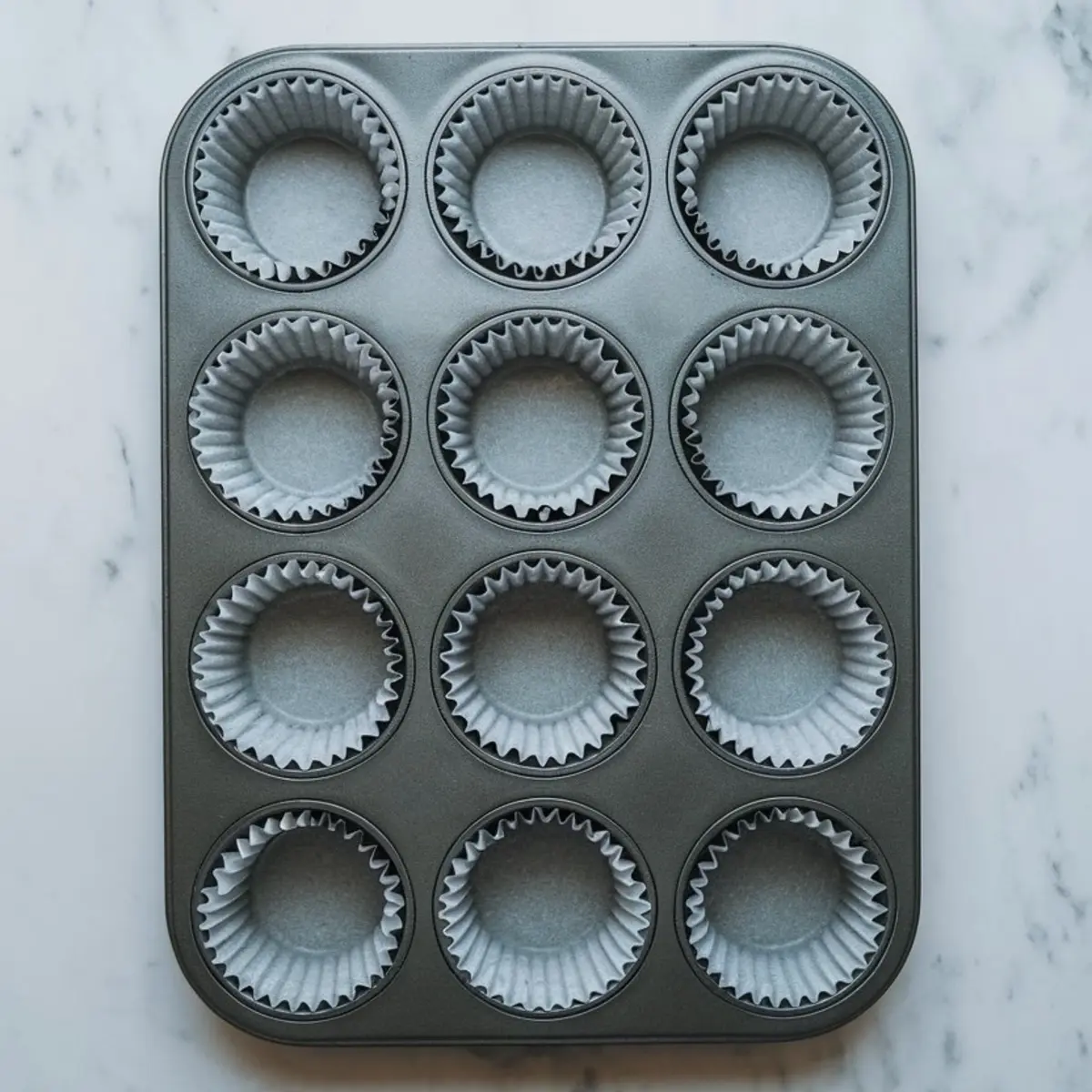 Empty nonstick muffin tin lined with white paper cupcake liners on a marble countertop, prepped for baking strawberry banana muffins.