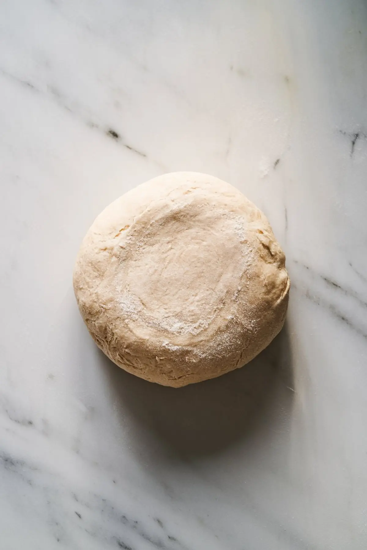 Overhead view of a round ball of cinnamon roll dough resting on a floured marble surface, ready for rolling and filling.