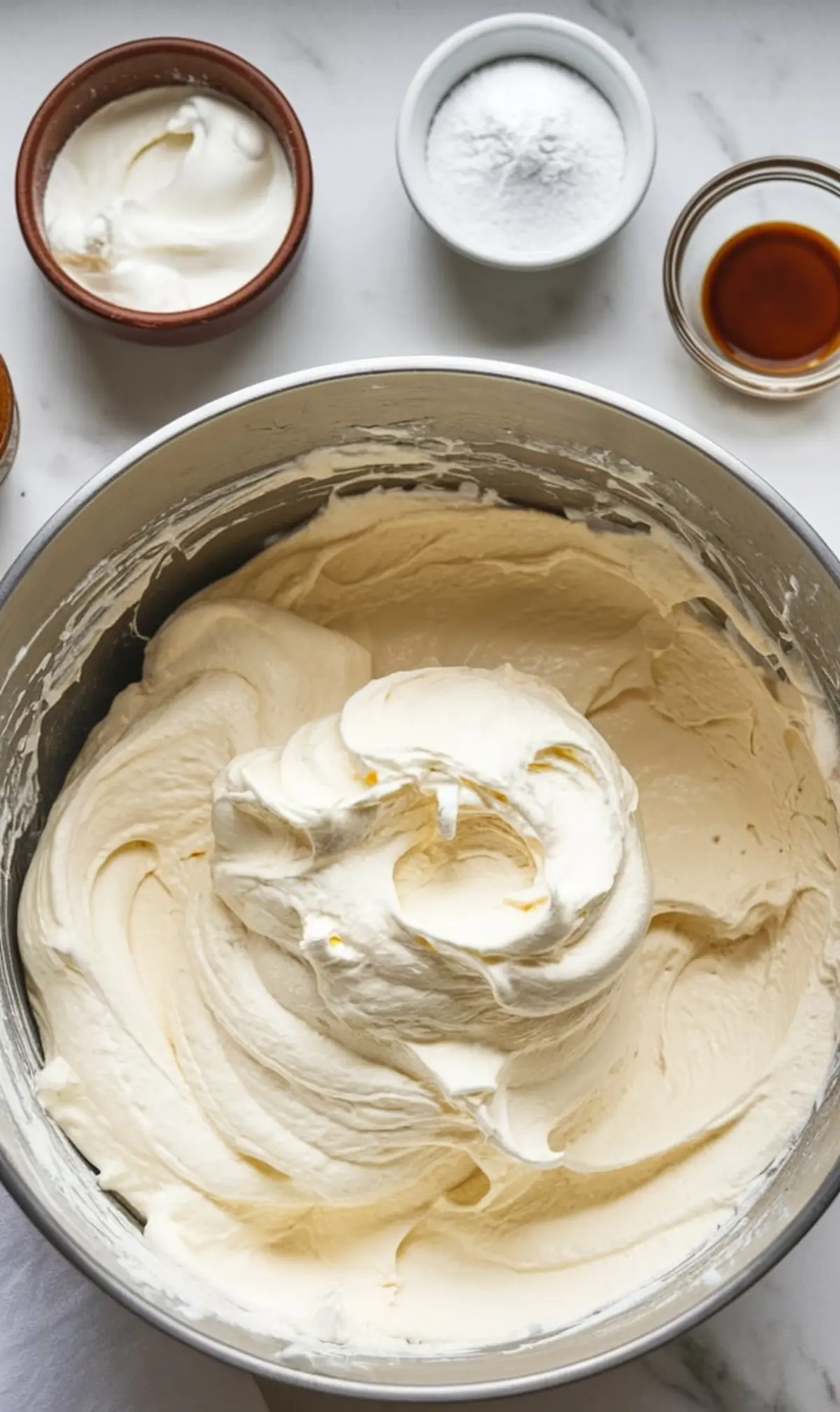Mixing bowl filled with thick whipped cream mixture surrounded by small bowls of vanilla extract, powdered sugar, and cream cheese on a white marble countertop, showing ingredients for homemade ice cream cake filling.